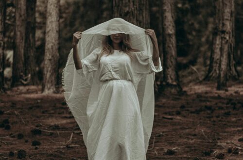 A woman in a white dress stands in a dark, eerie forest creating a mysterious atmosphere.