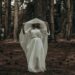 A woman in a white dress stands in a dark, eerie forest creating a mysterious atmosphere.