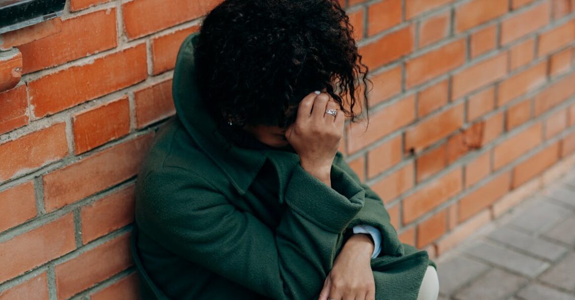 A woman in a green coat sits against a brick wall, deep in thought.