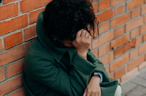 A woman in a green coat sits against a brick wall, deep in thought.