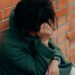 A woman in a green coat sits against a brick wall, deep in thought.